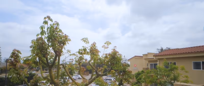 View of a senior living facility named Nikkei Senior Gardens with trees in the foreground, a parking lot with cars, and a beige building with a red-tiled roof under a partly cloudy sky.