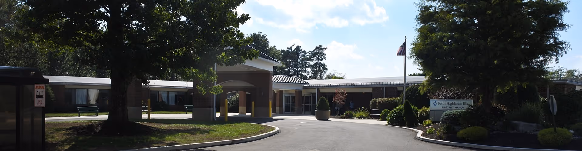 Exterior view of a single-story senior living facility building with a covered entrance, surrounded by trees and landscaping under a partly cloudy sky.