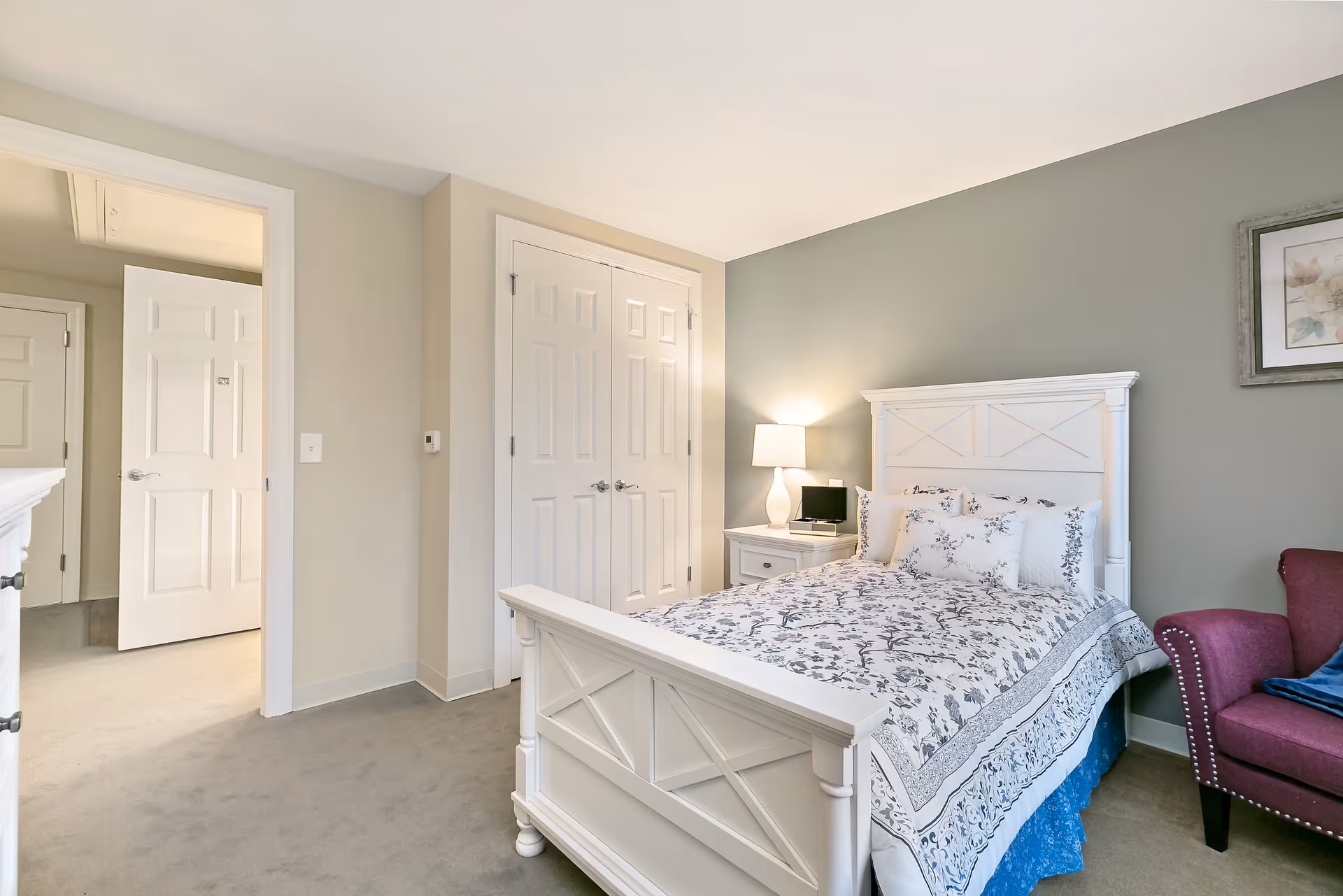 A bright bedroom with a white wooden bed frame and floral patterned bedding. There is a white nightstand with a lamp and a small black device on it. A maroon armchair with a blue cushion is positioned near the bed. The walls are painted light gray and beige, and there is a framed artwork hanging on the wall. Two white doors are visible, one open leading to another room and one closed closet door.
