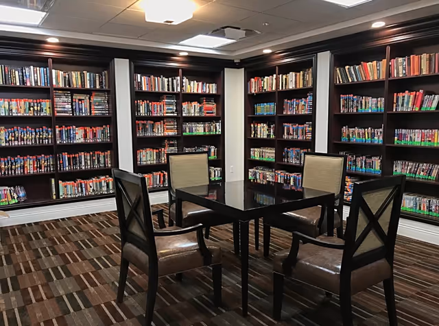 A cozy library room with dark wooden bookshelves filled with books lining the walls. In the center, there is a black square table surrounded by four cushioned chairs with wooden frames. The floor is carpeted with a patterned design, and the ceiling has recessed lighting.