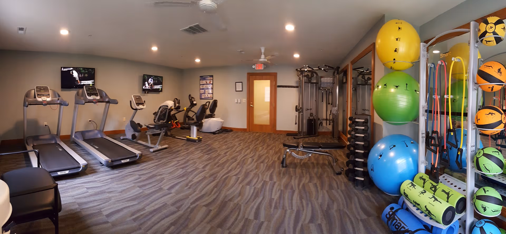 A fitness room with exercise equipment including two treadmills, two recumbent bikes, a multi-station gym machine, a bench, and a rack of dumbbells. On the right side, there are colorful exercise balls, resistance bands, and medicine balls organized on a rack. The room has carpeted flooring, beige walls, ceiling fans, and two wall-mounted televisions.