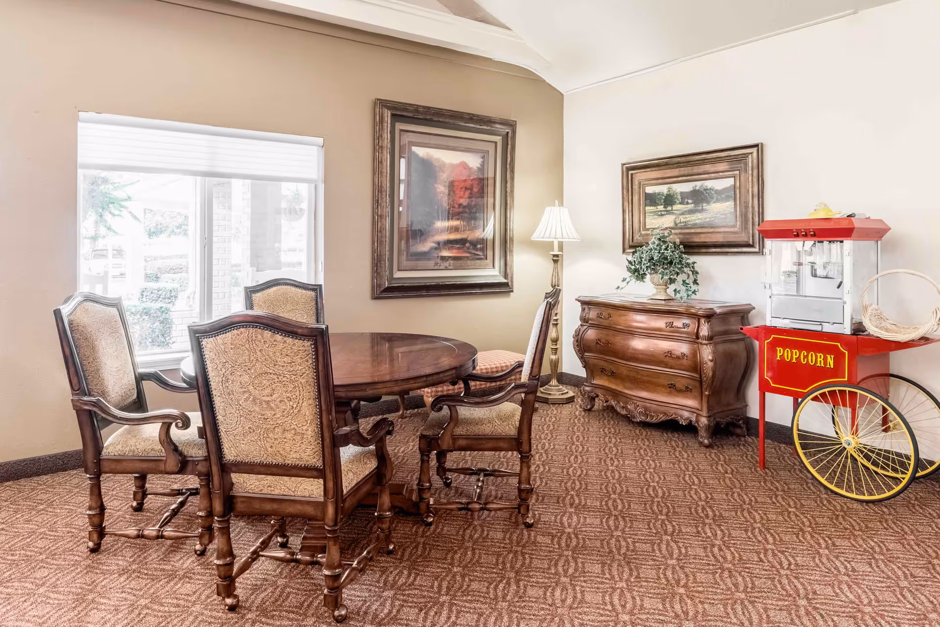 A cozy interior room with a round wooden table surrounded by four upholstered chairs. The room features a patterned carpet, two framed landscape paintings on the walls, a wooden dresser with a plant on top, a floor lamp, and a red popcorn machine on wheels.