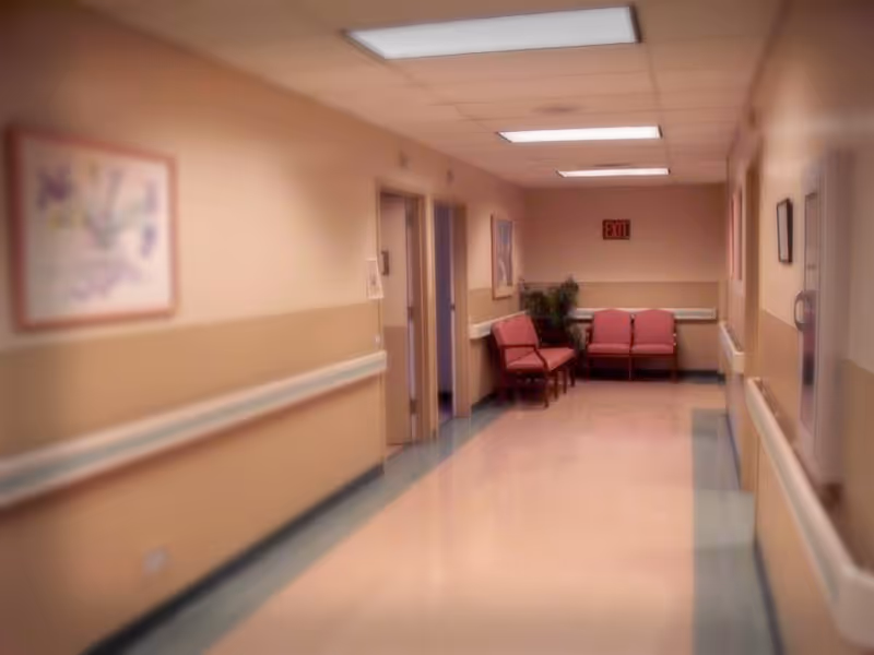 A softly lit hallway in a senior living facility with beige walls, handrails, and a tiled floor. There are several open doorways along the left side and a small seating area with red chairs and a potted plant at the end of the hallway. Artwork is hung on the walls and an exit sign is visible above the seating area.