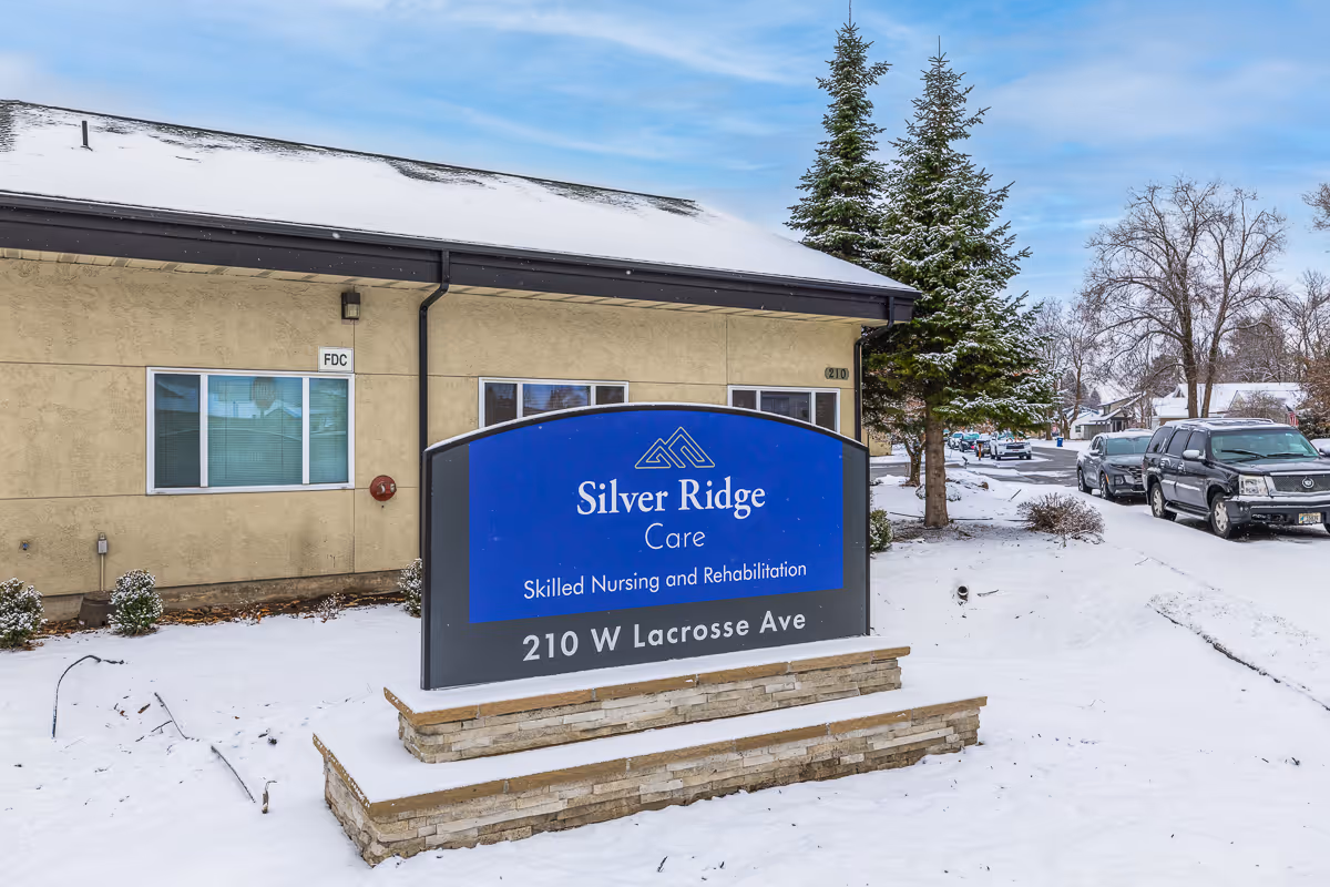 Snow-covered sign reading "Silver Ridge Care" in front of a beige building with parked cars on a snowy street.