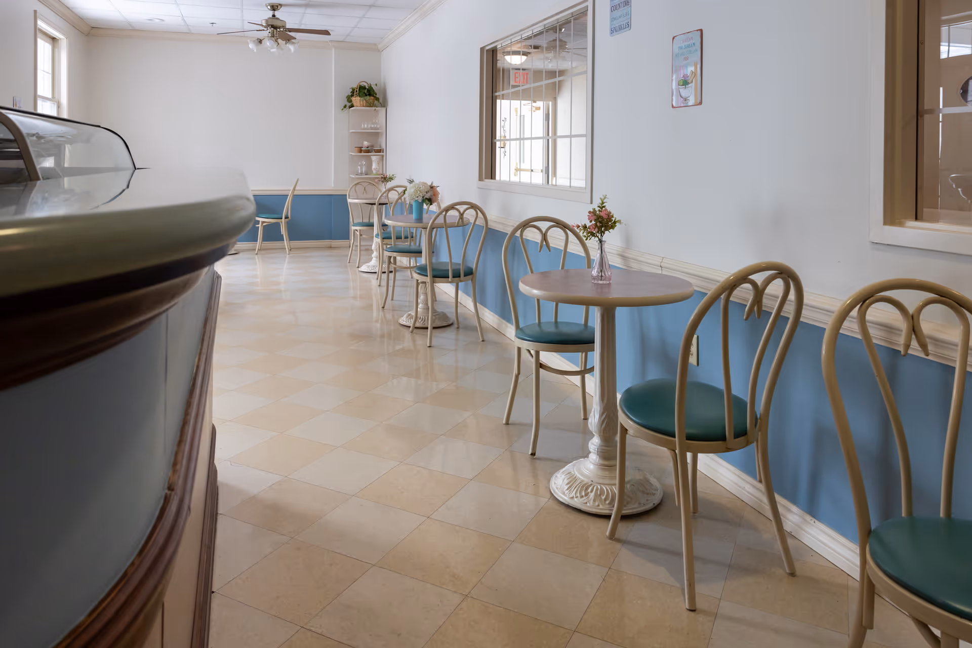 A small dining area with round tables and chairs lined up against a wall painted blue on the lower half and white on the upper half. Each table has a small vase with flowers. There is a counter on the left side and a ceiling fan above. The floor has a checkered pattern of beige tiles.