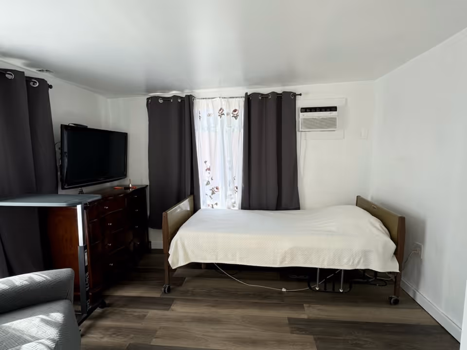 A small bedroom with a single bed covered in a white blanket, positioned against a white wall with an air conditioning unit above it. Next to the bed is a dark wooden dresser with a flat-screen TV mounted on the wall above it. The room has dark curtains on the windows and a light gray armchair partially visible in the foreground. The floor is covered with wood-patterned vinyl or laminate.
