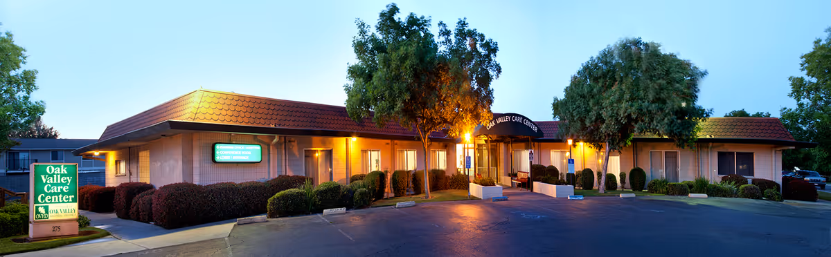 Front exterior of Oak Valley Care Center at dusk with an illuminated entrance, trees, landscaping, and a parking area.