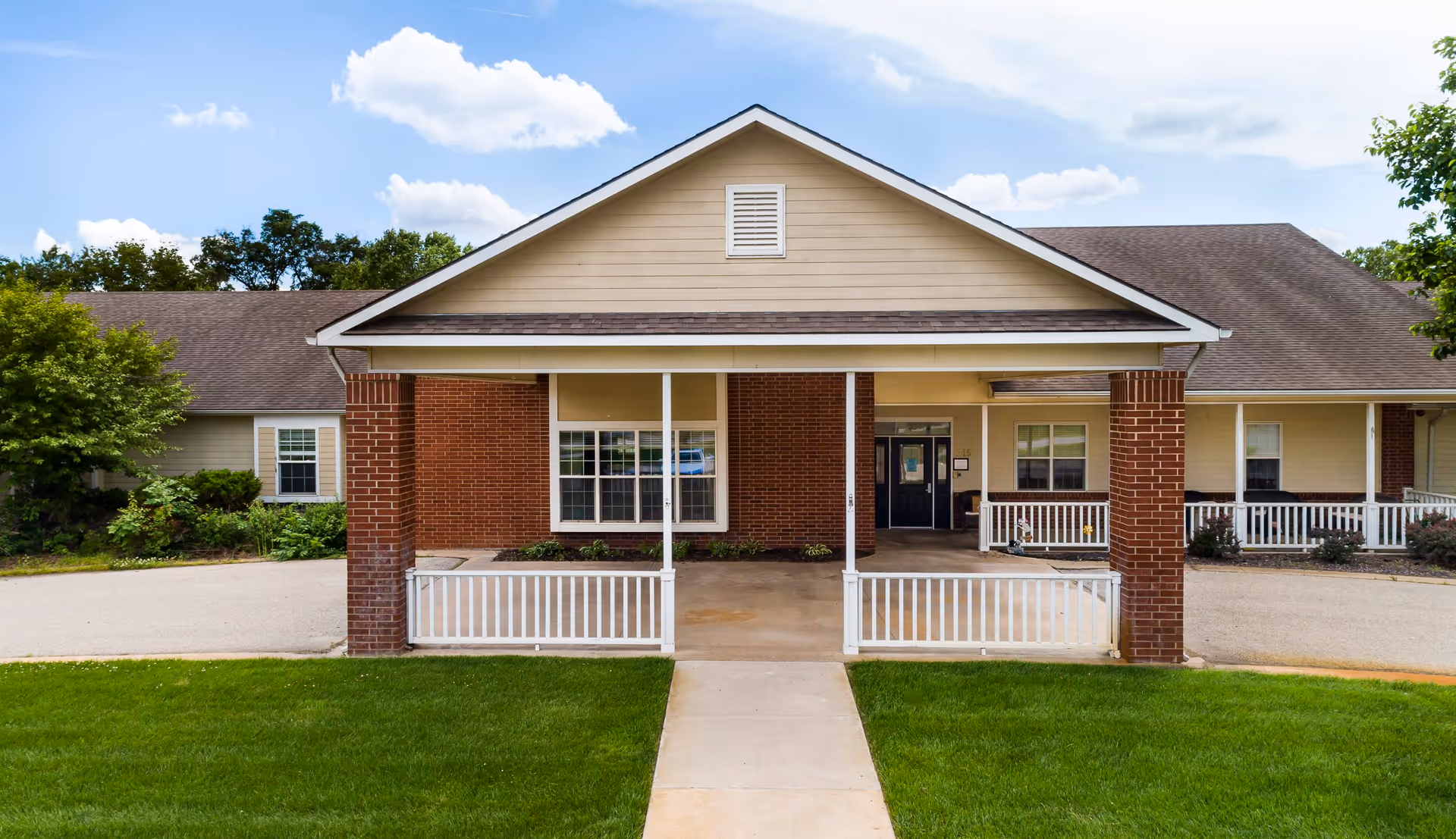 Front exterior view of Homestead Assisted Living of Halstead building with a covered entrance supported by brick columns, white railings, and a concrete walkway leading to the entrance. The building has beige siding and a dark shingled roof with windows on either side of the entrance.