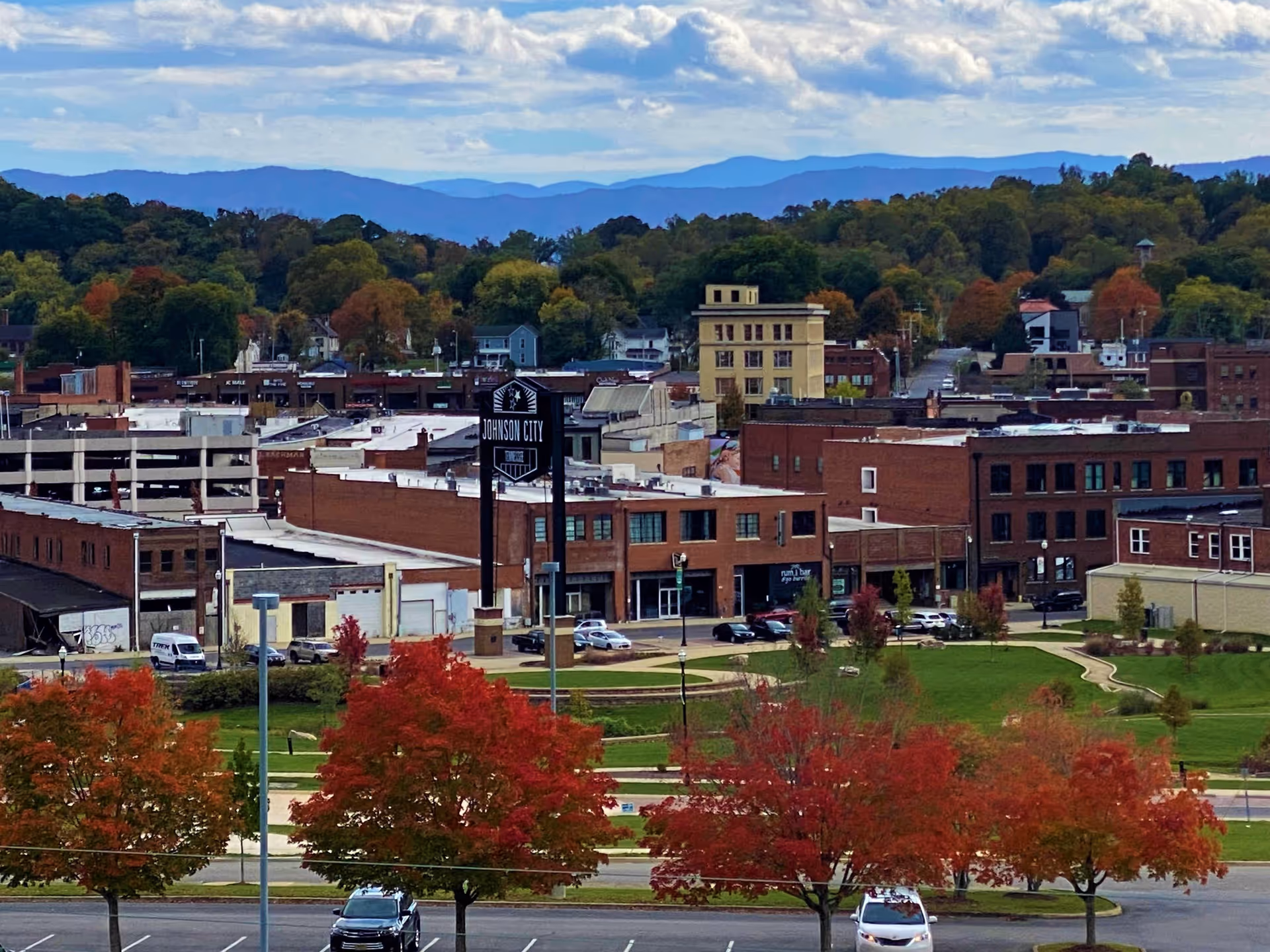 View of a small cityscape with brick buildings, a sign reading 'Johnson City', green park areas, and trees with autumn foliage in the foreground, with mountains visible in the background under a partly cloudy sky.