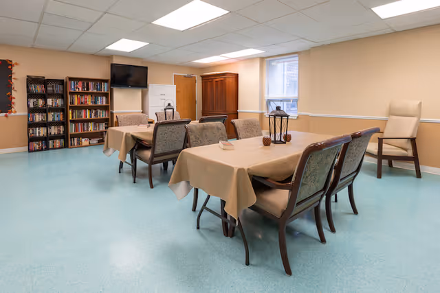 A well-lit room with two tables covered in beige tablecloths, each surrounded by cushioned chairs. The room has light blue flooring, beige walls, and a window letting in natural light. There are bookshelves filled with books and DVDs, a wall-mounted TV, a wooden cabinet, and a single armchair in the corner.