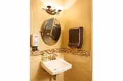 Small beige bathroom with a pedestal sink, round mirror, soap dispenser, wall-mounted paper towel dispenser, and a decorative mosaic tile border.