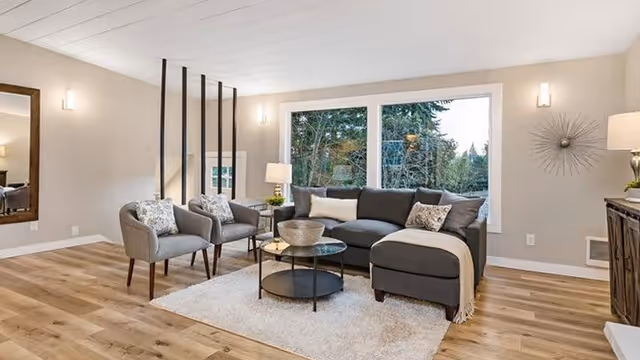 Bright modern living room with a gray sectional sofa, two armchairs, a round coffee table on a rug, and a large window overlooking trees.