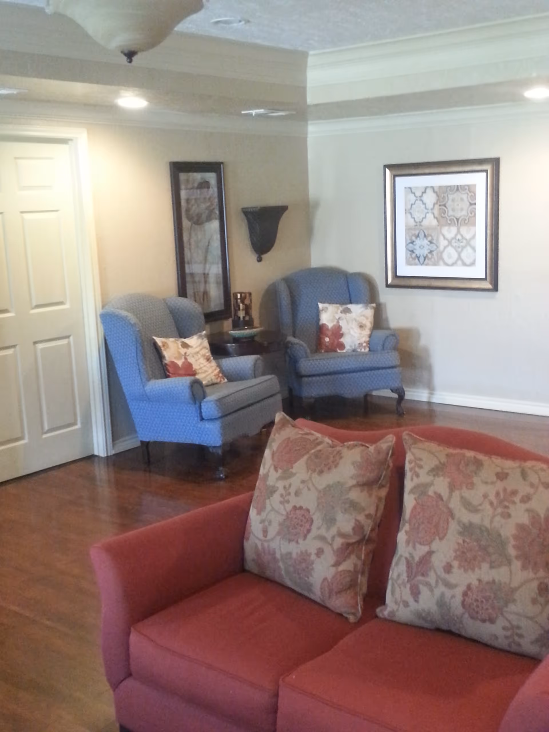 A cozy living room area with a red sofa adorned with two floral-patterned cushions in the foreground. In the background, there are two blue upholstered armchairs, each with a decorative pillow, positioned around a small round wooden table. The walls are beige with two framed artworks and a wall-mounted sconce light fixture. The floor is wooden, and a closed white door is visible on the left side.