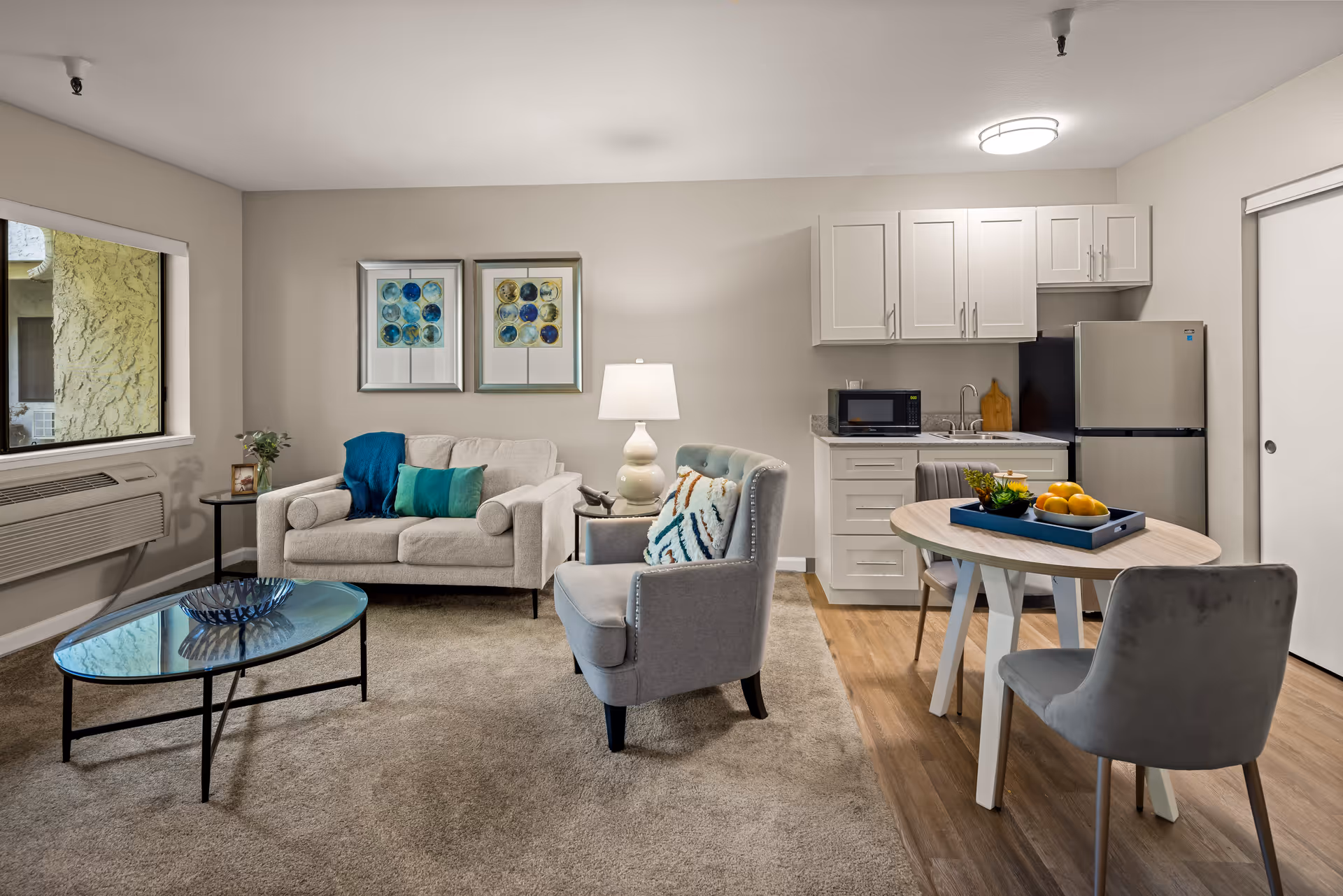 A cozy senior living room and kitchenette area featuring a beige loveseat with blue and green pillows, a gray armchair with a patterned cushion, a glass coffee table, and a small round dining table with two gray chairs. The kitchenette includes white cabinets, a microwave, a sink, and a refrigerator. Two framed artworks hang on the wall above the loveseat, and a window with an air conditioning unit is on the left side.