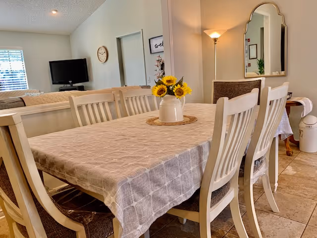 A dining area with a rectangular table covered by a beige checkered tablecloth and six white wooden chairs. A white vase with sunflowers is placed in the center of the table. In the background, there is a living room area with a TV mounted on the wall, a clock, a floor lamp, a mirror, and a small table with a white cloth.