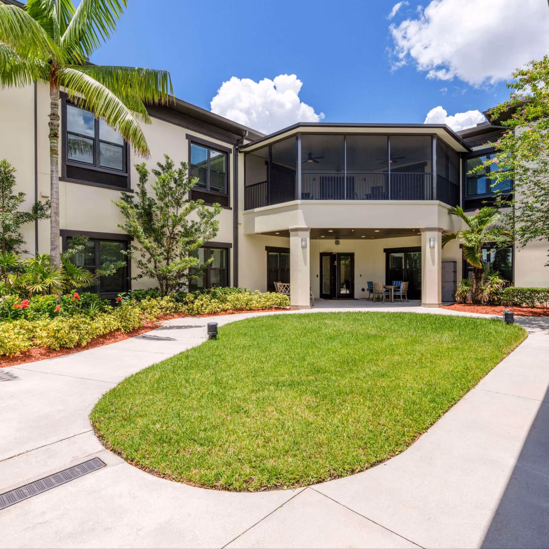 Outdoor courtyard area of Arbor Terrace Cooper City featuring a well-maintained green lawn surrounded by a concrete walkway, tropical plants, and palm trees. The two-story building has large windows and a screened balcony on the upper floor. There is outdoor seating under the balcony on the ground floor, and the sky is clear with some clouds.