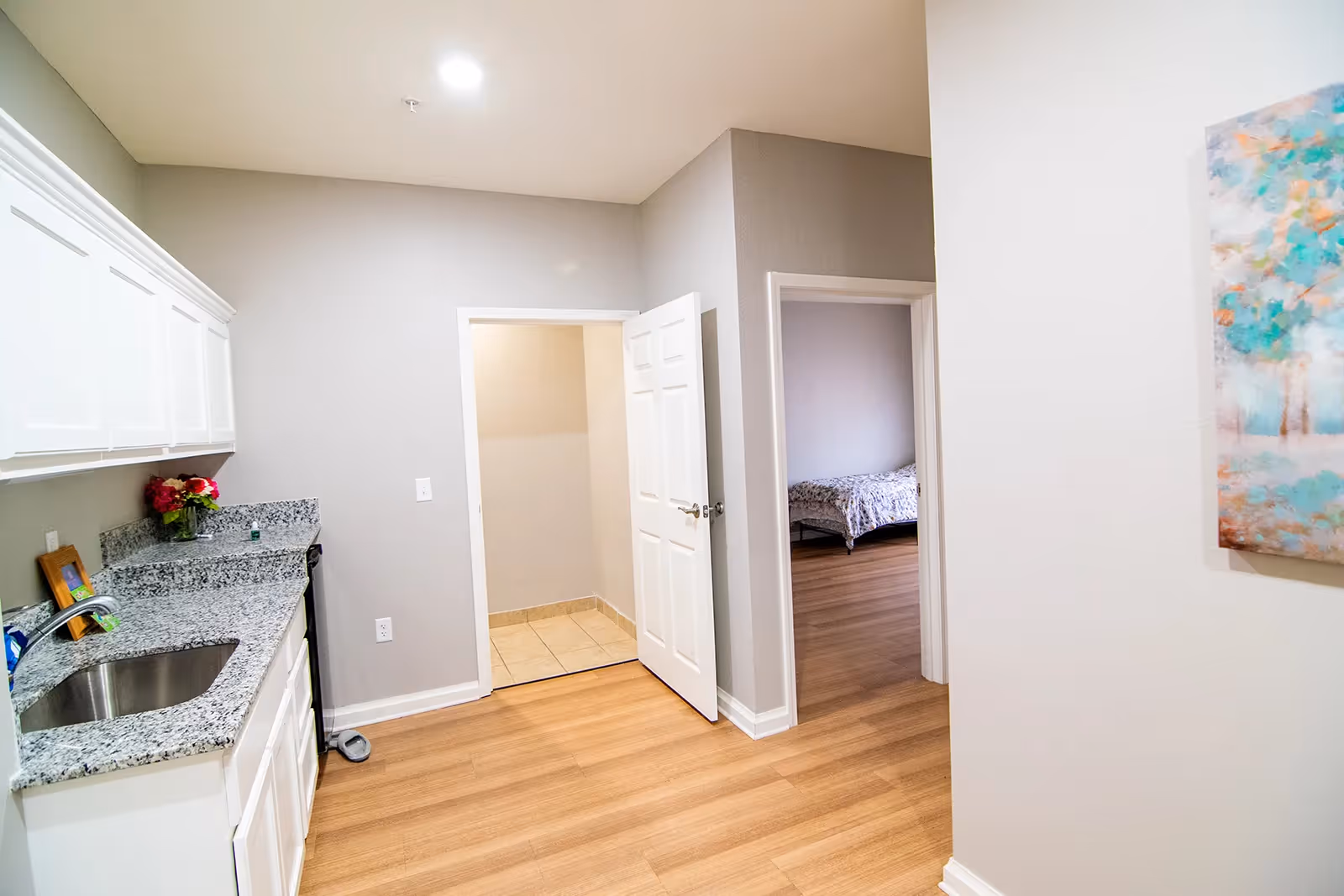 Interior view of a kitchen area with white cabinets and granite countertops on the left side. There is a stainless steel sink and a vase with flowers on the counter. The floor is wooden, and there are two doorways visible: one leading to a tiled room and another leading to a bedroom with a bed covered in a floral bedspread. A colorful abstract painting is partially visible on the right wall.