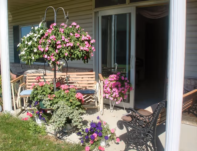 A patio area with hanging flower baskets and potted plants in front of a sliding glass door. There is a wooden bench with cushions and a decorative black metal chair on the concrete patio, adjacent to a grassy lawn.