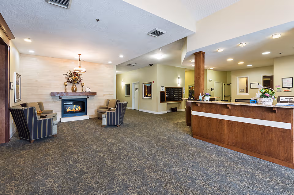 A spacious assisted living facility lobby with a wooden reception desk on the right, a seating area with four armchairs arranged around a modern fireplace on the left, and decorative flowers on the mantel. The walls are light-colored, and the carpet has a subtle pattern. The area is well-lit with ceiling lights.