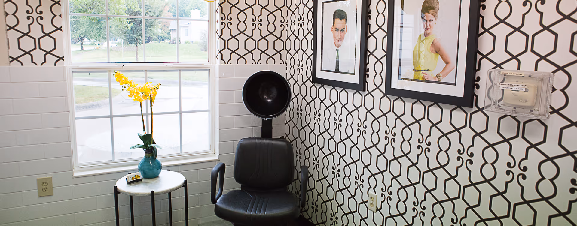 A small room with a black salon hair dryer chair positioned against a wall with black and white geometric patterned wallpaper. Next to the chair is a small round table with a blue vase holding yellow flowers. Two framed portraits hang on the wall above the chair. A window with a view of green trees and a street is on the left side of the room.