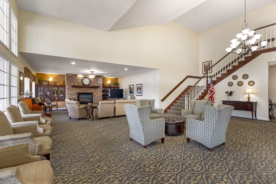 A spacious living room area with patterned carpet, several upholstered armchairs arranged around a round ottoman, and a seating area with sofas and chairs near a brick fireplace with a clock above it. There is a large window on the left side letting in natural light, a staircase with wooden railing on the right, and decorative plates on the wall next to a small table with a lamp and flowers.