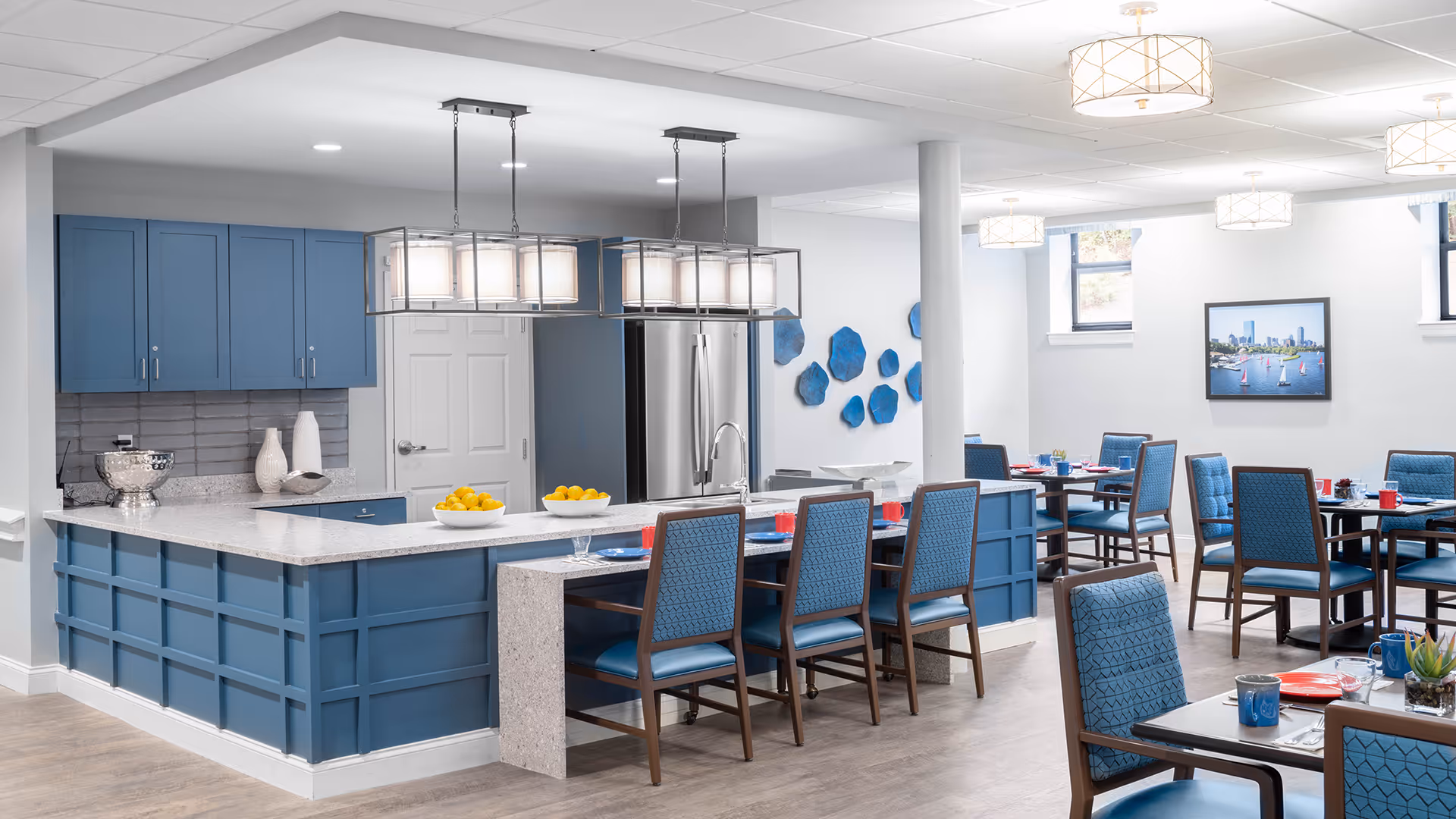 A modern dining area with a kitchen island and multiple dining tables. The kitchen features blue cabinetry, a white countertop, stainless steel refrigerator, and pendant lighting. The dining tables have blue cushioned chairs and are set with plates, cups, and utensils. The room has light-colored walls, wood flooring, and decorative blue wall art. A framed picture of a city skyline with sailboats is on the wall.