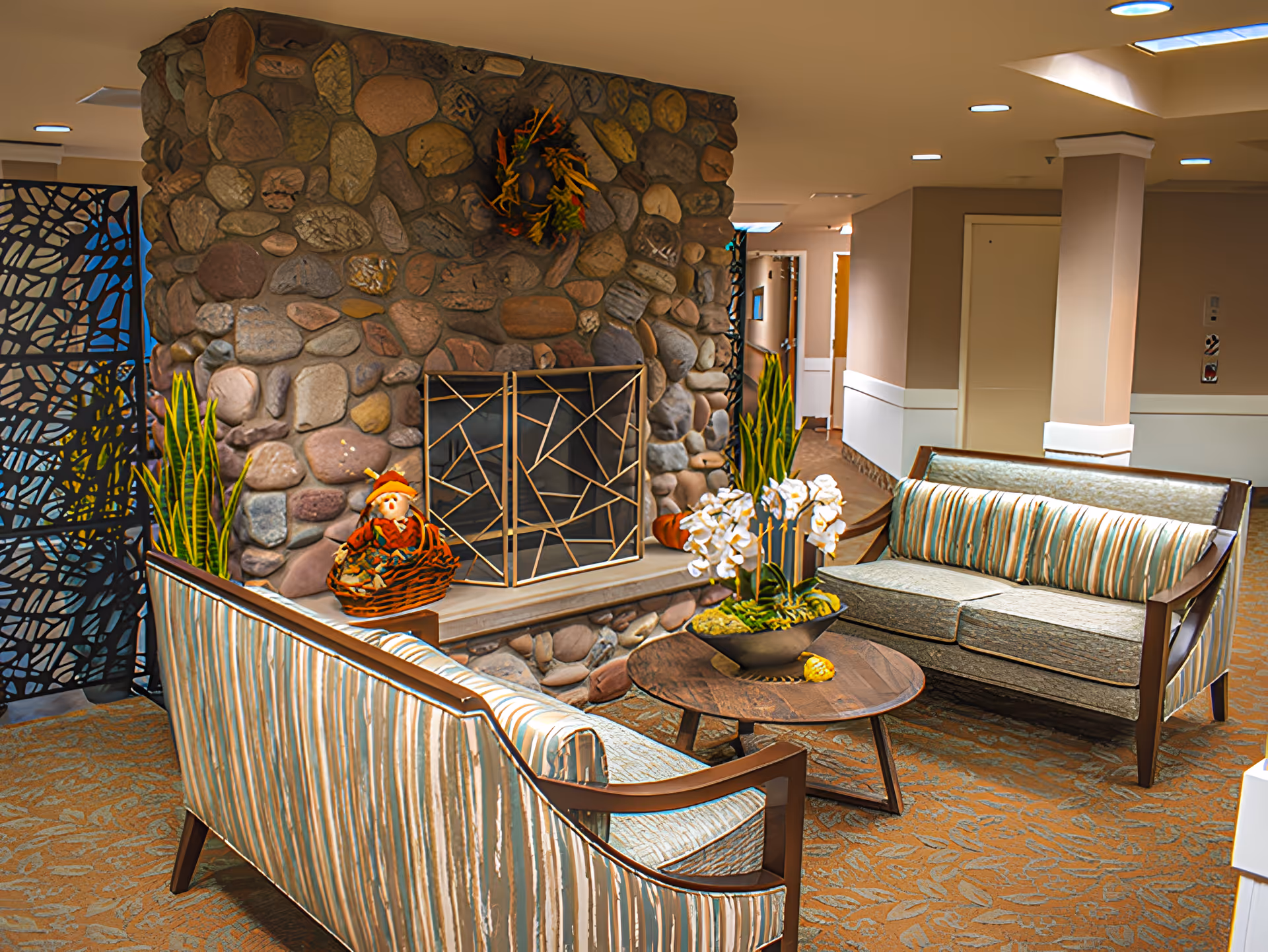 A cozy lounge with striped sofas, a round wooden coffee table topped with orchids, and a stone fireplace in a senior living facility.
