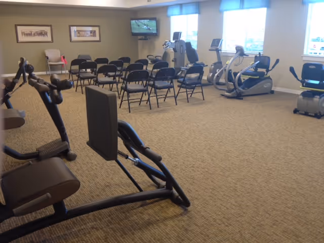 A fitness/activity room with exercise machines and rows of folding chairs facing a wall-mounted TV under windows.