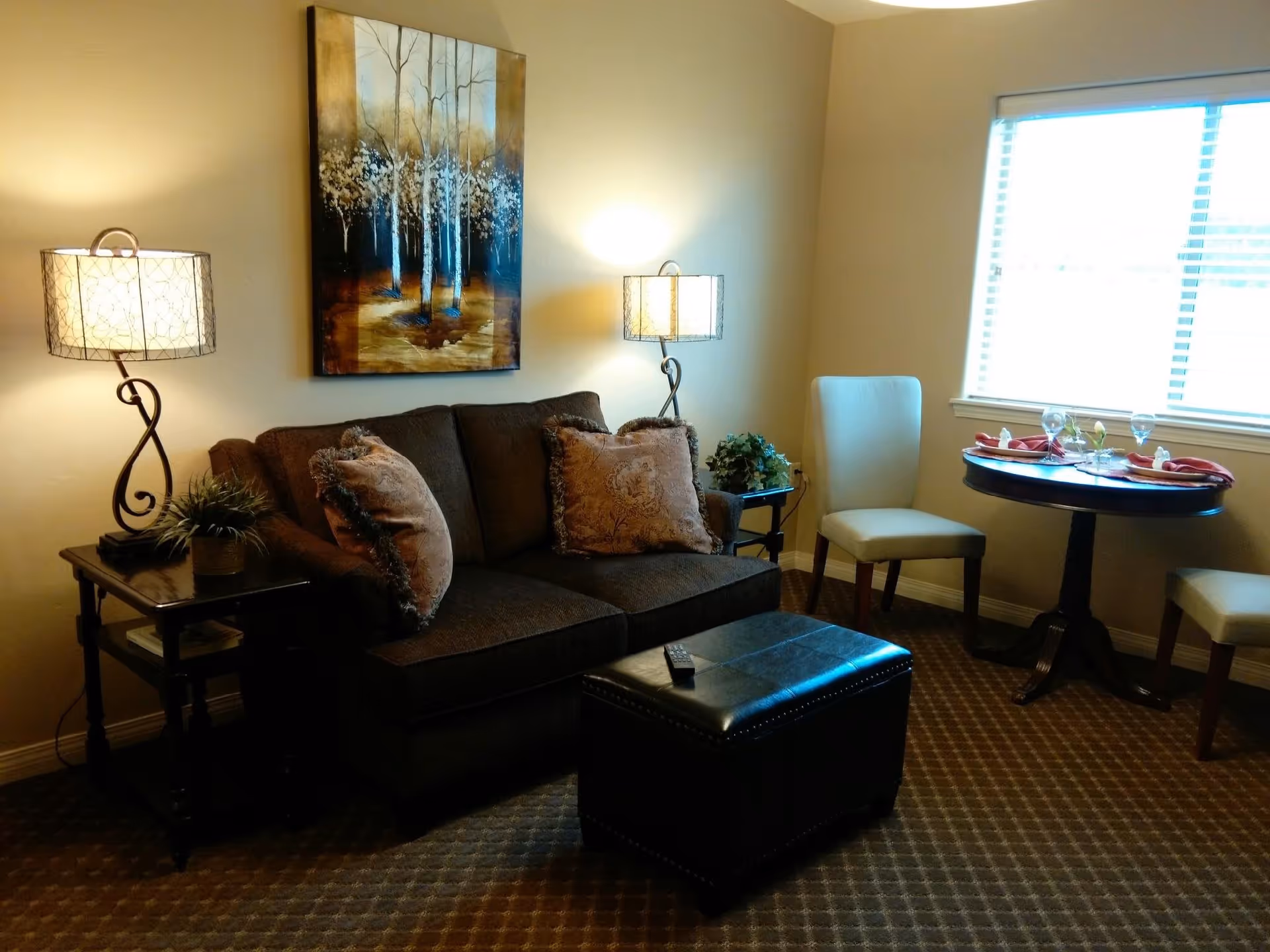 A cozy living room area with a dark brown sofa adorned with two decorative pillows, flanked by two side tables each holding a lamp with a patterned shade. Above the sofa hangs a painting of trees. In front of the sofa is a black leather ottoman with a remote control on top. To the right, there is a small round dining table set with two chairs, place settings, and napkins, positioned near a window with blinds letting in natural light.