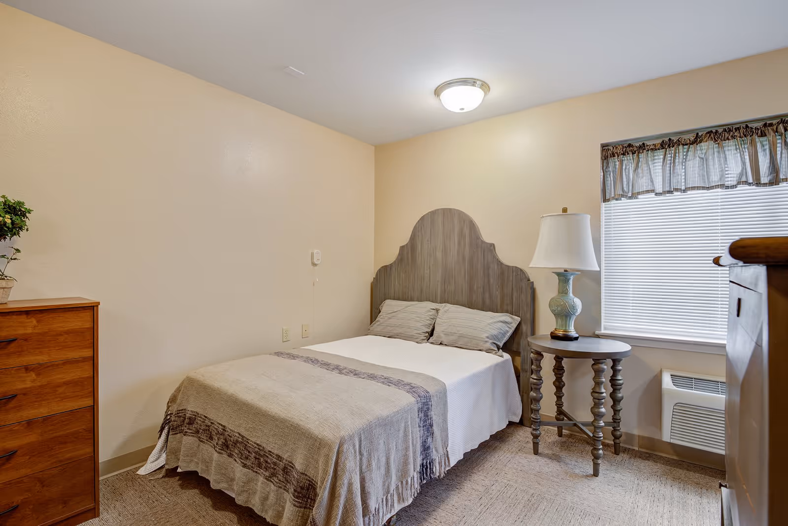A small, neatly arranged bedroom with a double bed featuring a wooden headboard, two pillows, and a beige blanket with a decorative stripe. To the right of the bed is a round wooden side table with a decorative lamp. A window with blinds and a valance is on the right wall. On the left side of the room is a wooden dresser with a small potted plant on top. The walls are painted a light beige color, and the floor is carpeted.