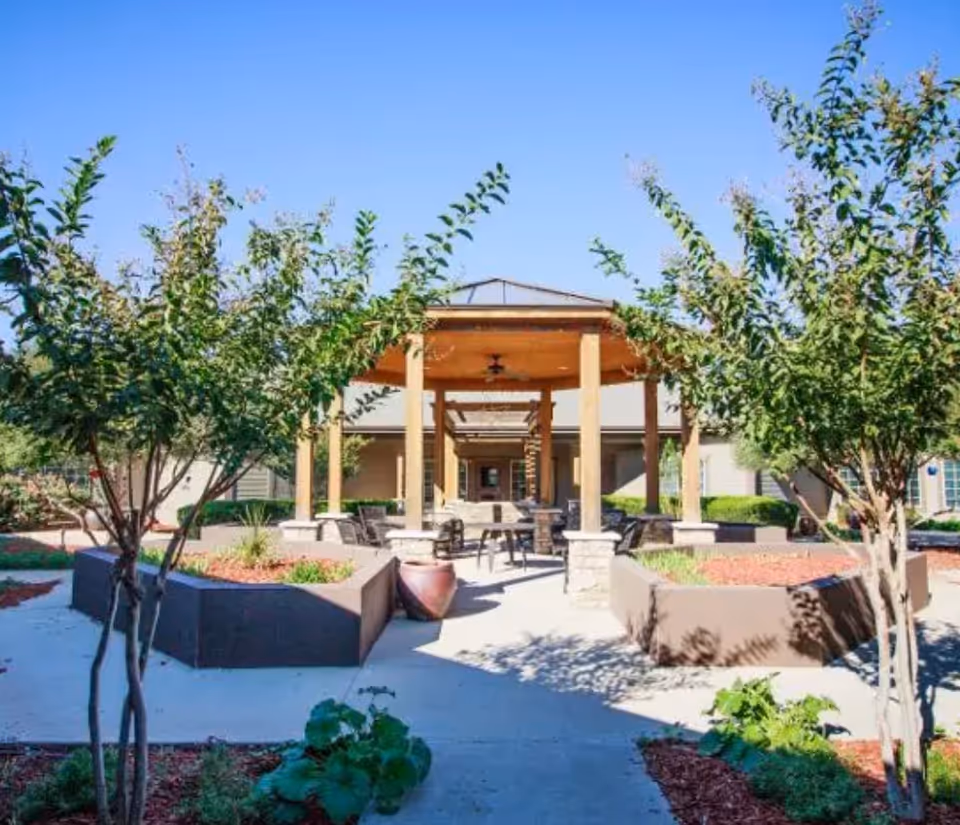 Outdoor seating area at Legacy Ranch featuring a wooden pergola with ceiling fans, surrounded by landscaped garden beds with small trees and plants under a clear blue sky.