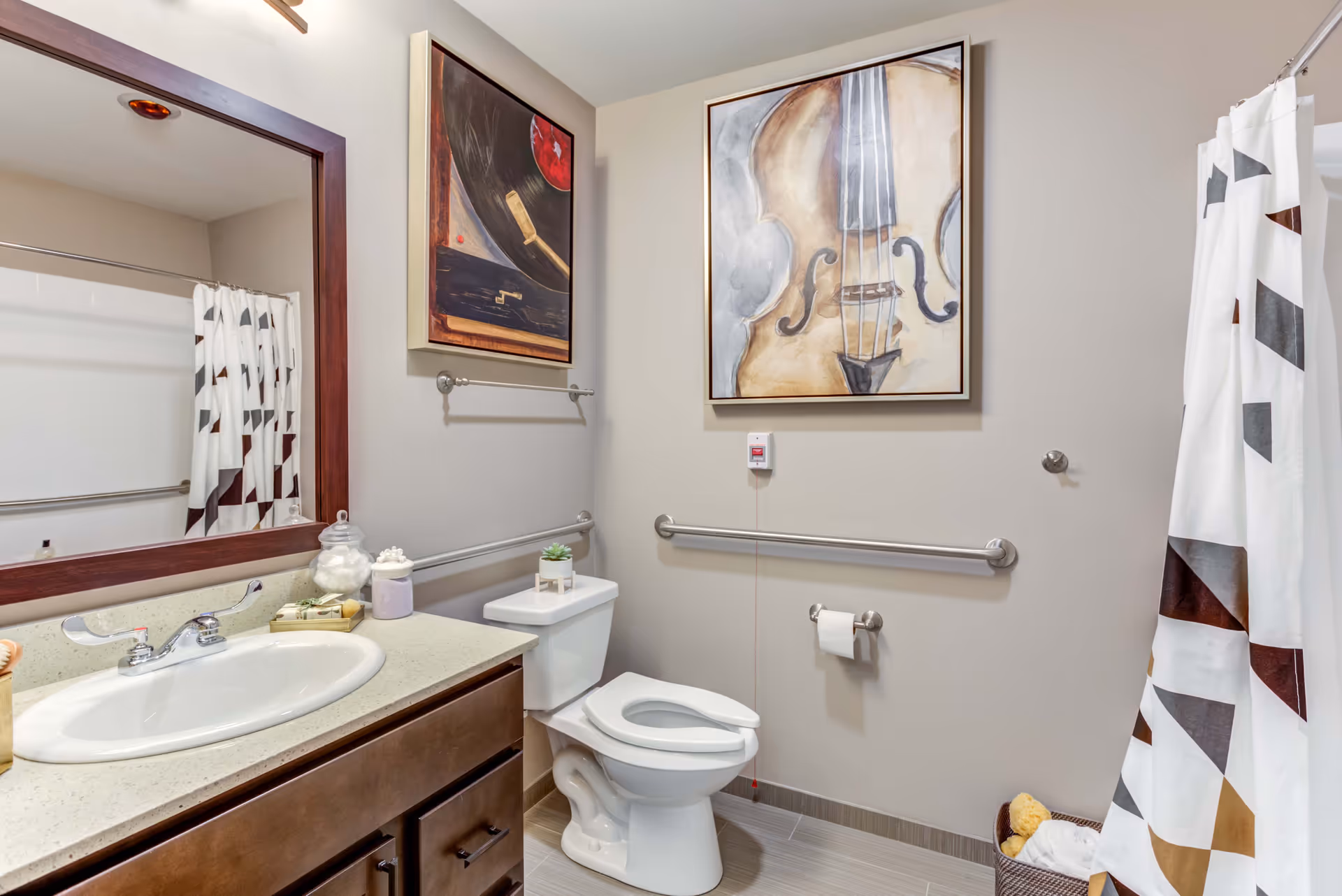 A clean and accessible bathroom with a white toilet, a sink with a countertop, and a large mirror above the sink. There are two framed artworks on the walls, one depicting a violin and the other a close-up of a record player. The bathroom has grab bars installed near the toilet and bathtub, and a shower curtain with a geometric pattern. A basket with towels and a sponge is placed on the floor near the bathtub.