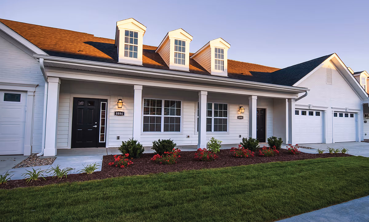 Exterior view of a residential building with white siding, black doors, and three dormer windows on the roof. There are two entrances with house numbers 8894 and 8899, outdoor wall lanterns, a well-maintained garden with red flowers and green shrubs, and a neatly trimmed lawn in front.