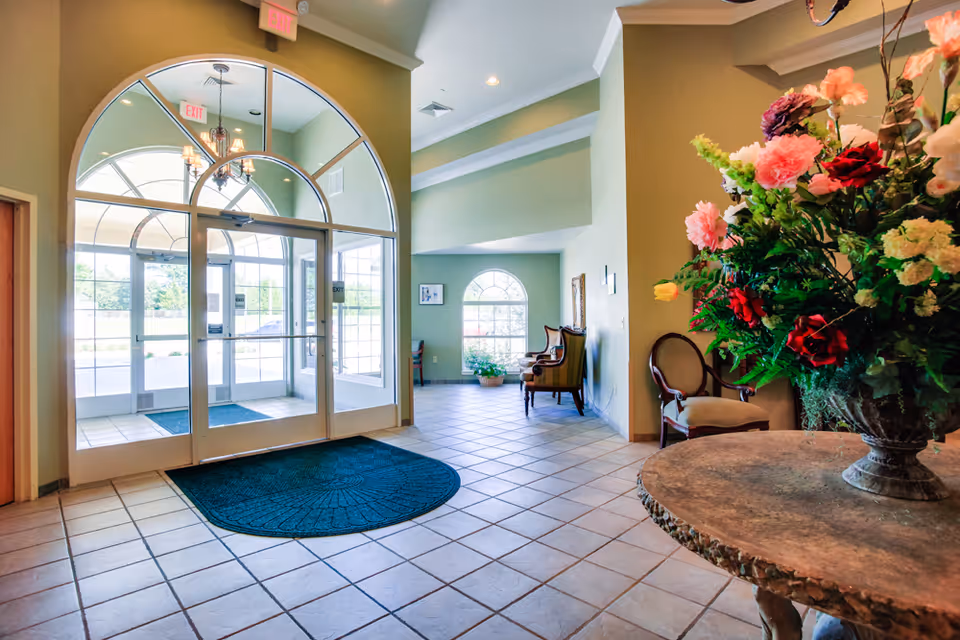 Bright and spacious entrance lobby of a senior living facility with large glass double doors featuring arched windows above. The floor is tiled, and there is a dark blue mat in front of the doors. To the right, a round stone table holds a large vase filled with colorful flowers. In the background, there are several chairs arranged near a window with an arched top, allowing natural light to fill the space.