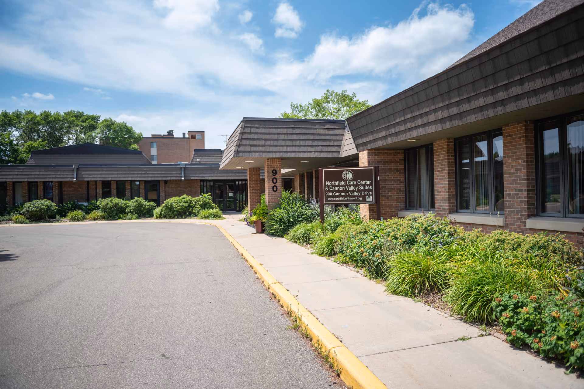 Exterior view of a single-story brick building with a brown shingled roof, a sidewalk, and landscaped greenery along the front. A sign near the entrance reads 'Northfield Care Center & Cannon Valley Suites, 900 Cannon Valley Drive'. The sky is partly cloudy.