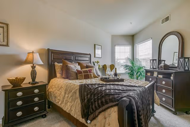 A cozy bedroom with a large wooden bed adorned with multiple pillows and a beige quilt. There is a dark brown throw blanket at the foot of the bed. To the left of the bed is a nightstand with a decorative lamp and a bowl. On the right side of the room is a wooden dresser with a large mirror and decorative items on top. Two windows with white blinds allow natural light into the room, and a green potted plant is placed near the corner.
