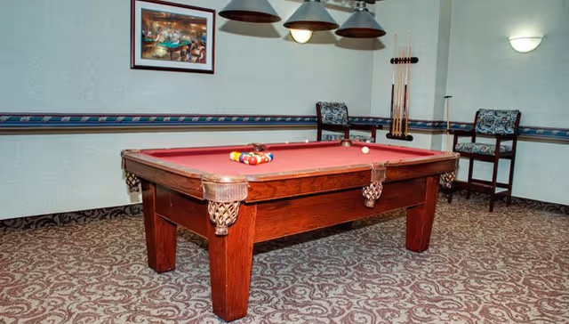 A room with a red felt pool table set up for a game, with pool balls racked and a cue ball on the table. Two patterned chairs are placed against the wall, which has a decorative border and a framed picture. Three hanging lamps are positioned above the pool table, and a wall-mounted light fixture is visible on the right side.