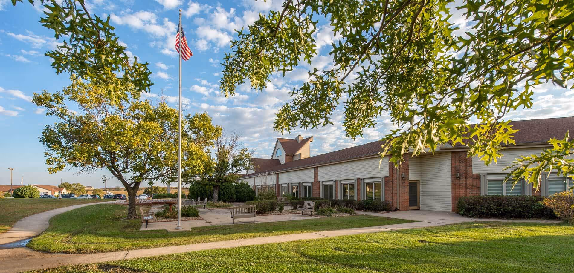 Exterior view of Emporia Presbyterian Manor showing a single-story building with large windows, a flagpole with an American flag, benches, trees, and a well-maintained lawn under a partly cloudy sky.