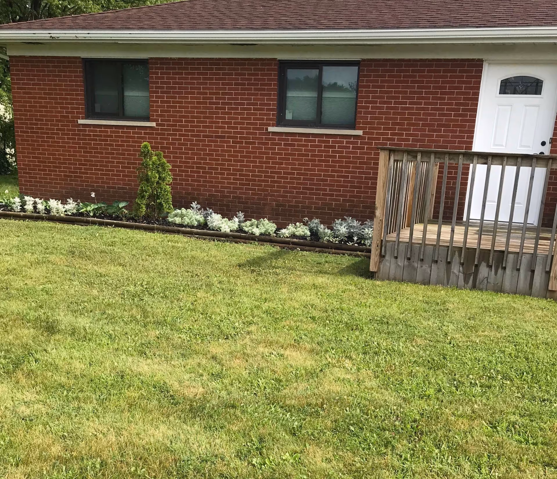 A red brick building with two windows and a white door with a small wooden porch and railing. In front of the building is a well-maintained green lawn and a flower bed with small plants and shrubs.