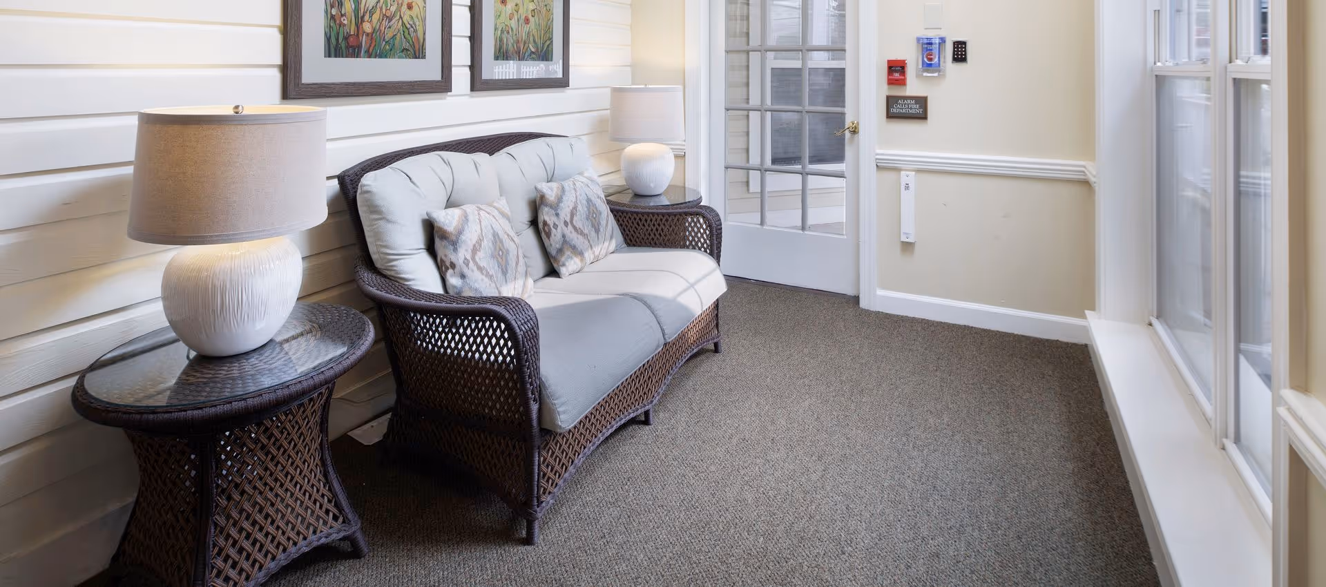 A cozy indoor seating area with a cushioned wicker loveseat and matching side tables, each holding a white lamp. The walls are light-colored with two framed floral artworks hanging above the loveseat. A glass door and large windows let in natural light, creating a bright and inviting atmosphere.