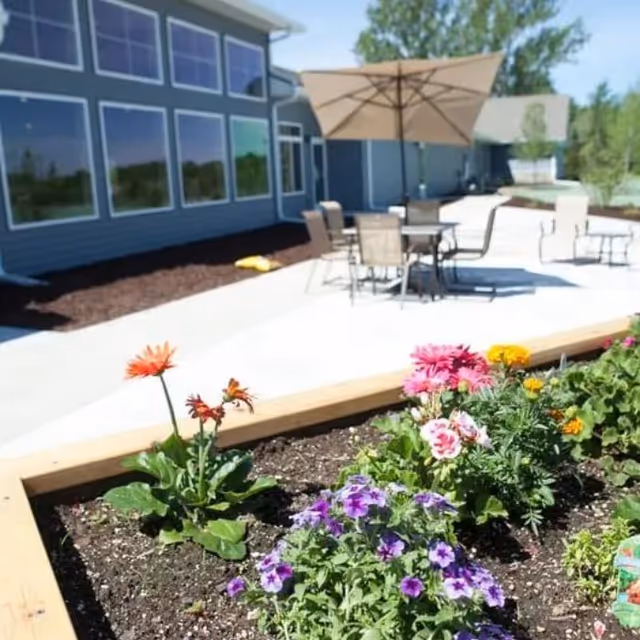 Outdoor patio area with a table, chairs, and a large umbrella next to a building with multiple windows. In the foreground, there is a raised garden bed with colorful flowers including purple, pink, orange, and yellow blooms.