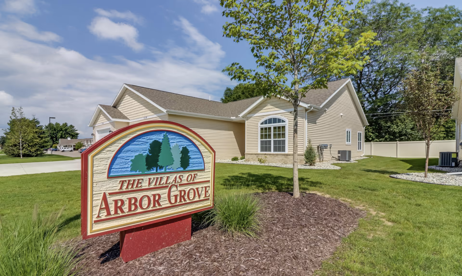Exterior view of a beige single-story building with a gable roof, surrounded by green grass, small trees, and landscaping. In the foreground, there is a large sign that reads 'The Villas of Arbor Grove' with an illustration of trees above the text. The sky is partly cloudy.