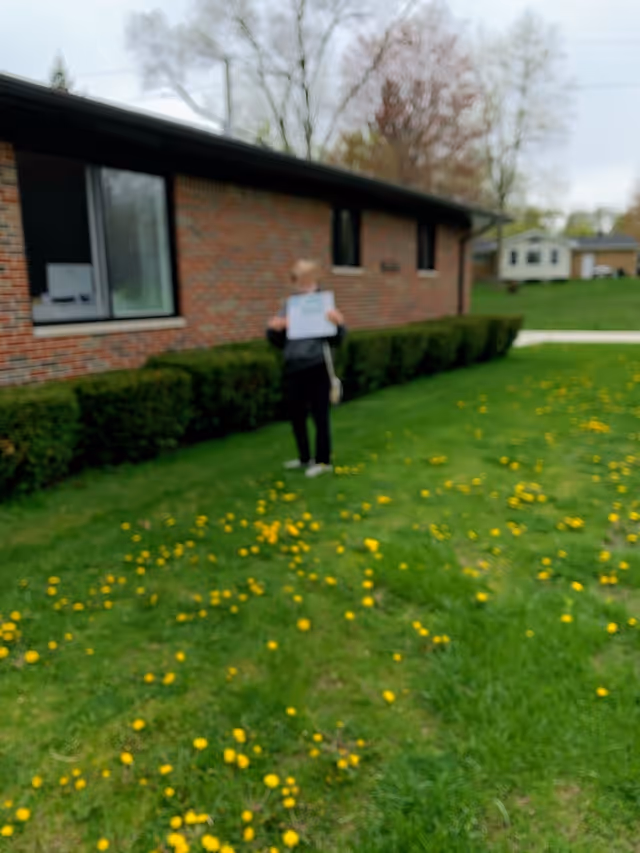 A person standing on a grassy lawn with yellow flowers in front of a brick building with windows. The person is holding a sign, but the image is blurry and details are unclear.