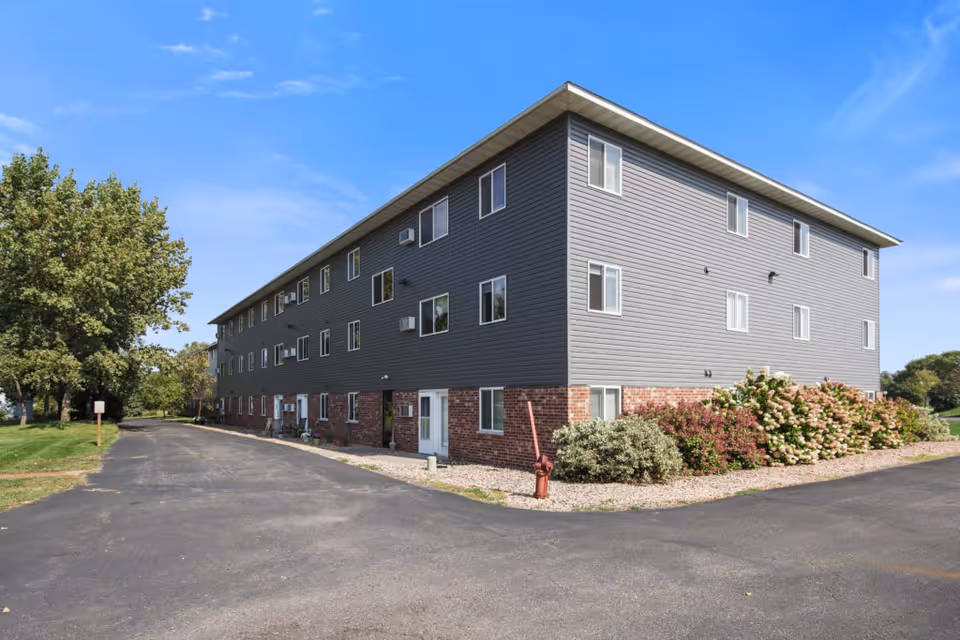 Exterior view of a three-story senior living facility building with gray siding and a brick base, surrounded by greenery and a paved driveway under a clear blue sky.