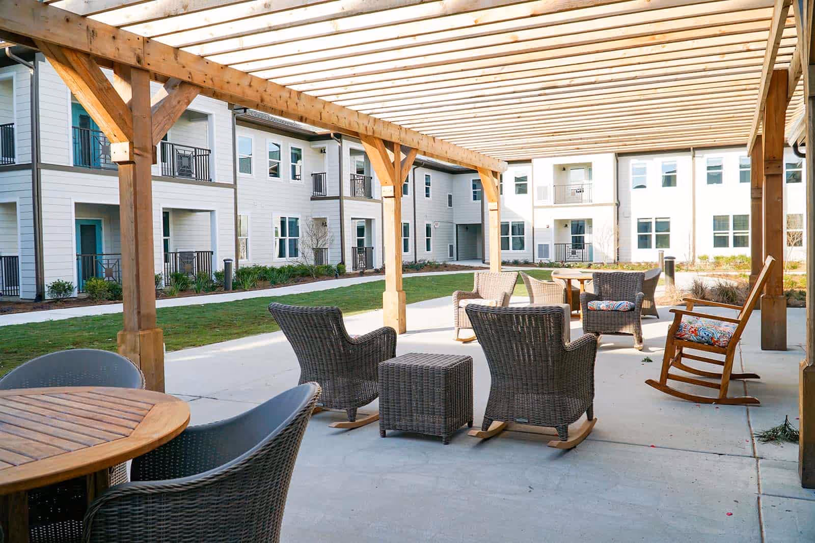 Outdoor covered patio area with wooden pergola, wicker rocking chairs and armchairs with cushions, round wooden tables, and a view of a two-story white building with balconies and windows in the background.
