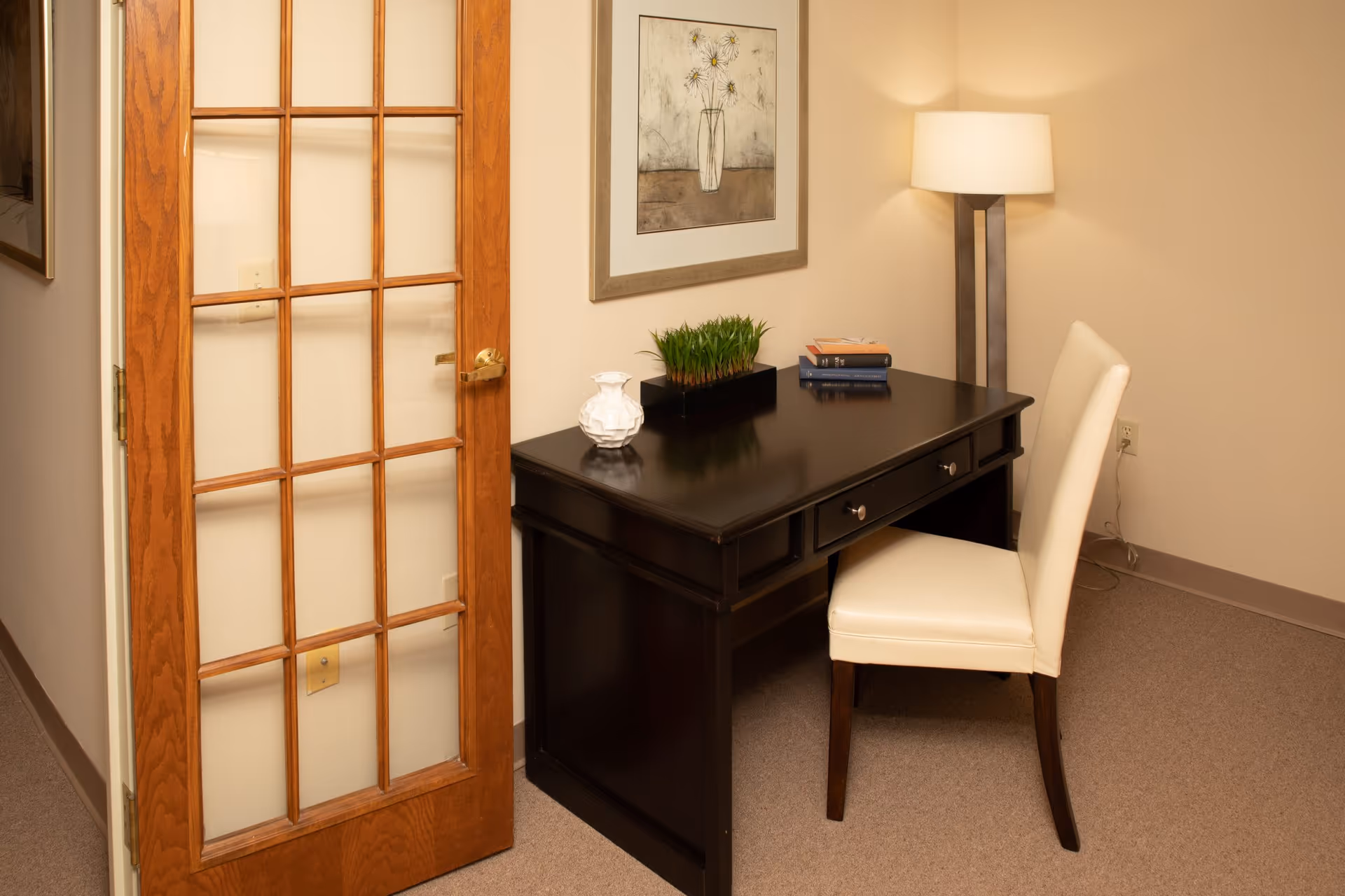 A small office or study area with a dark wooden desk, a white cushioned chair, a floor lamp, and a framed floral artwork on the wall. The room has beige walls and carpet, and a wooden door with glass panes is partially open.
