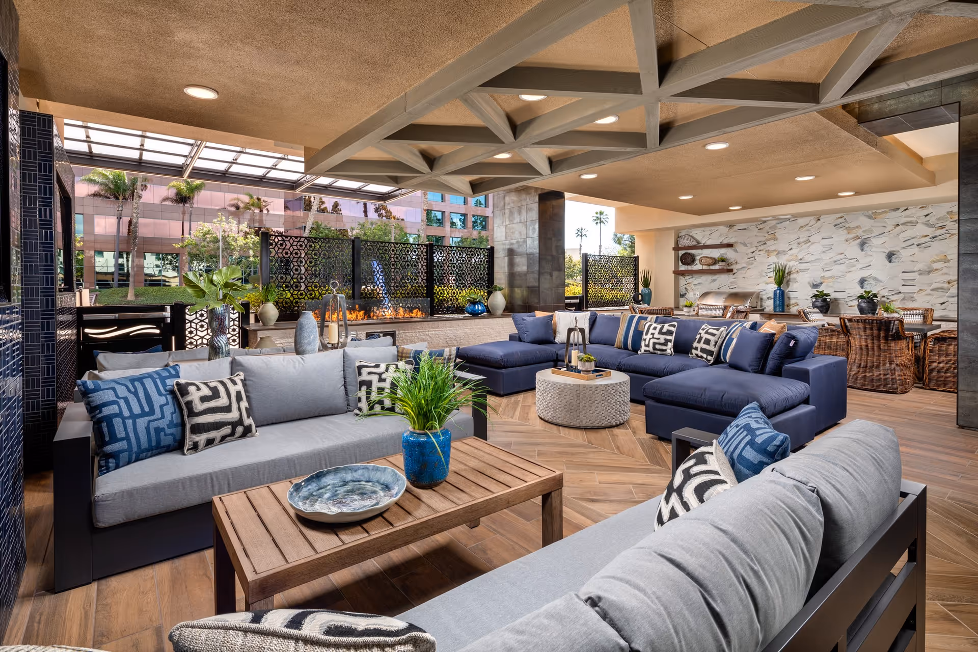 A spacious and stylish outdoor lounge area with modern sofas in gray and navy blue, decorated with patterned pillows. There is a wooden coffee table with a blue vase and decorative plate. The area features a decorative fire pit with a patterned screen behind it, and a dining table with wicker chairs in the background. The ceiling has exposed beams and recessed lighting, and large windows reveal palm trees and a building outside.