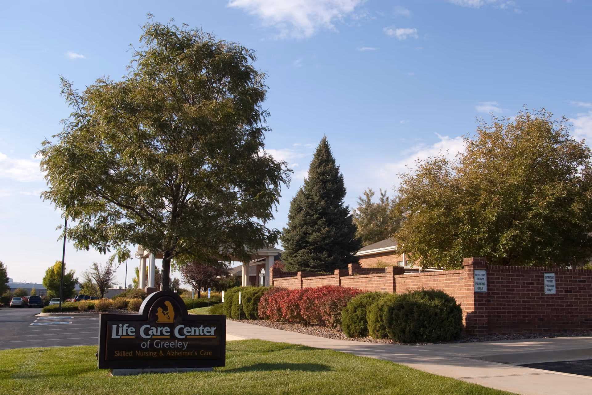 Exterior view of Life Care Center of Greeley showing a landscaped area with trees, bushes, a brick wall, and a sign indicating skilled nursing and Alzheimer's care. The sky is clear with some clouds.