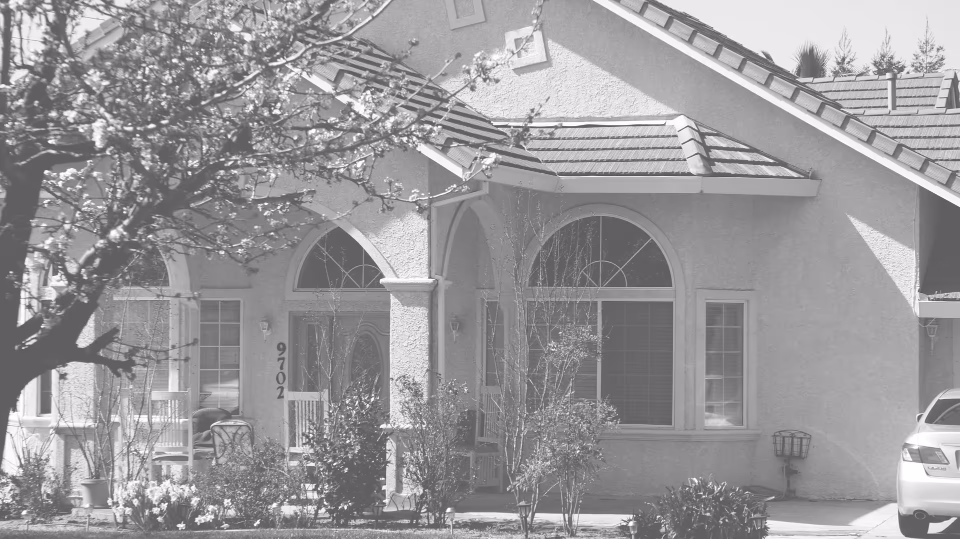 Front façade of a single-story stucco house with arched windows, a small porch with plants, and a parked car.