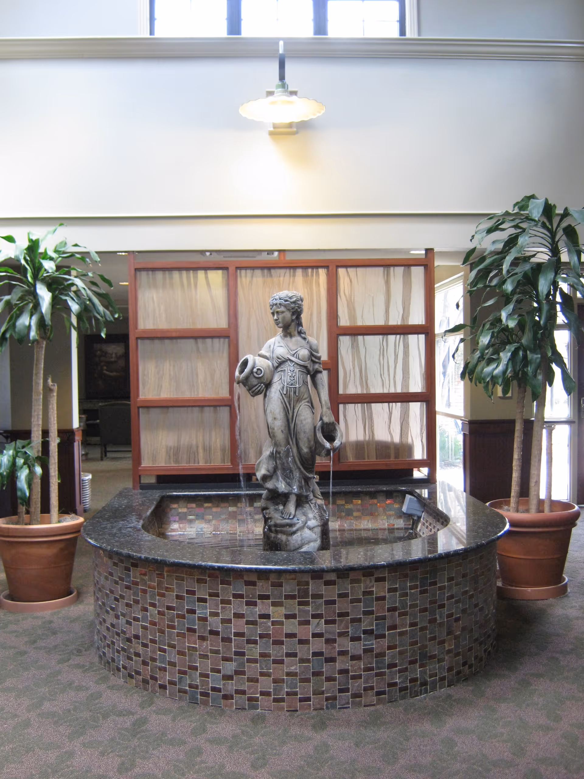 Indoor decorative water fountain with a statue of a woman holding two jugs, placed in a tiled circular basin. The fountain is flanked by two potted plants with tall green leaves. Behind the fountain is a wooden framed partition with translucent panels, and above is a hanging light fixture.